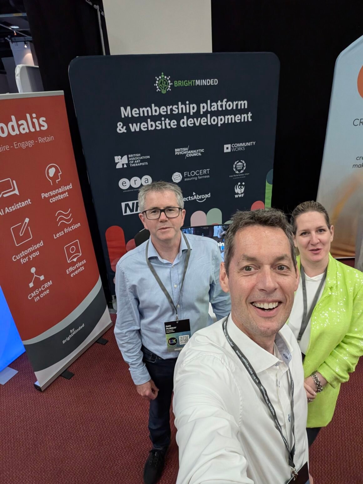 Three Caucasian people (two men and a woman) are taking a selfie infront of their exhibition stand in the background 